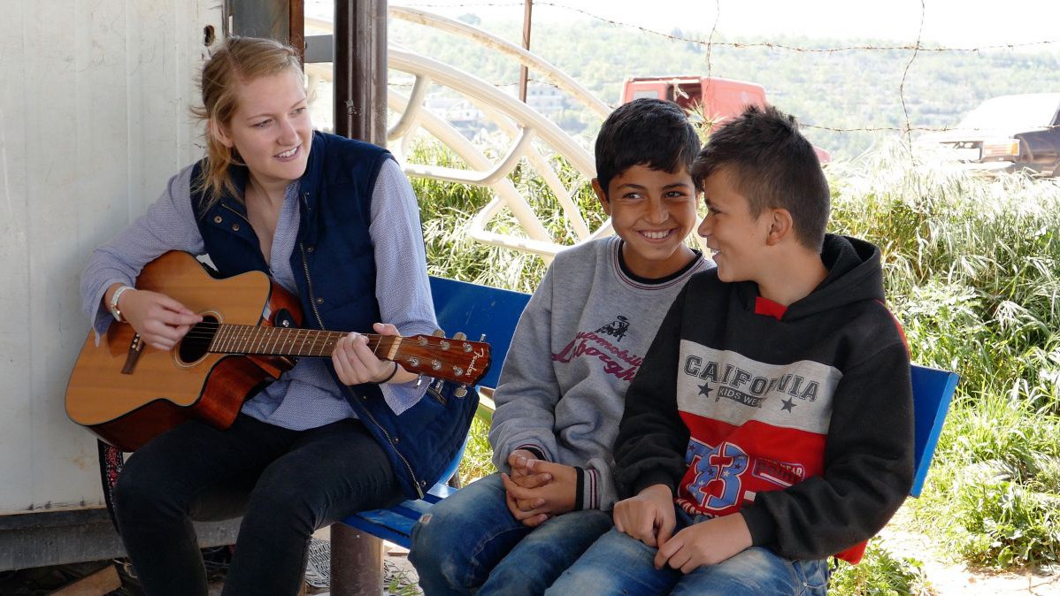 Zwei Kinder sitzen auf einer Band gemeinsam mit einer Frau, die Gitarre spielt.