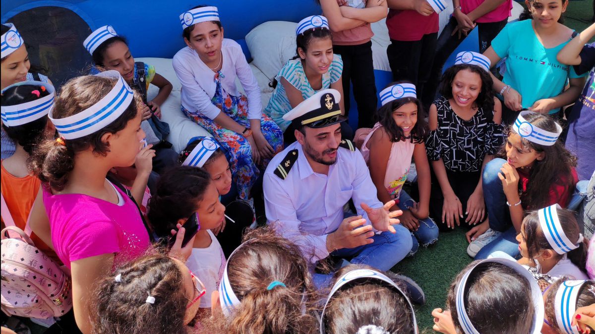 Ein Mann als Kapitän verkleidet sitzt am Boden, um ihn herum viele Kinder mit Matrosenmützen.