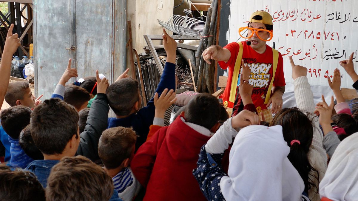 Billy Cookie schenkt Kindern in einem Flüchtlingslager eine unbeschwerte Zeit. Der Clown Billy Cookie inmitten einer Gruppe von Kindern, die mit ihrer Hand aufzeigen.