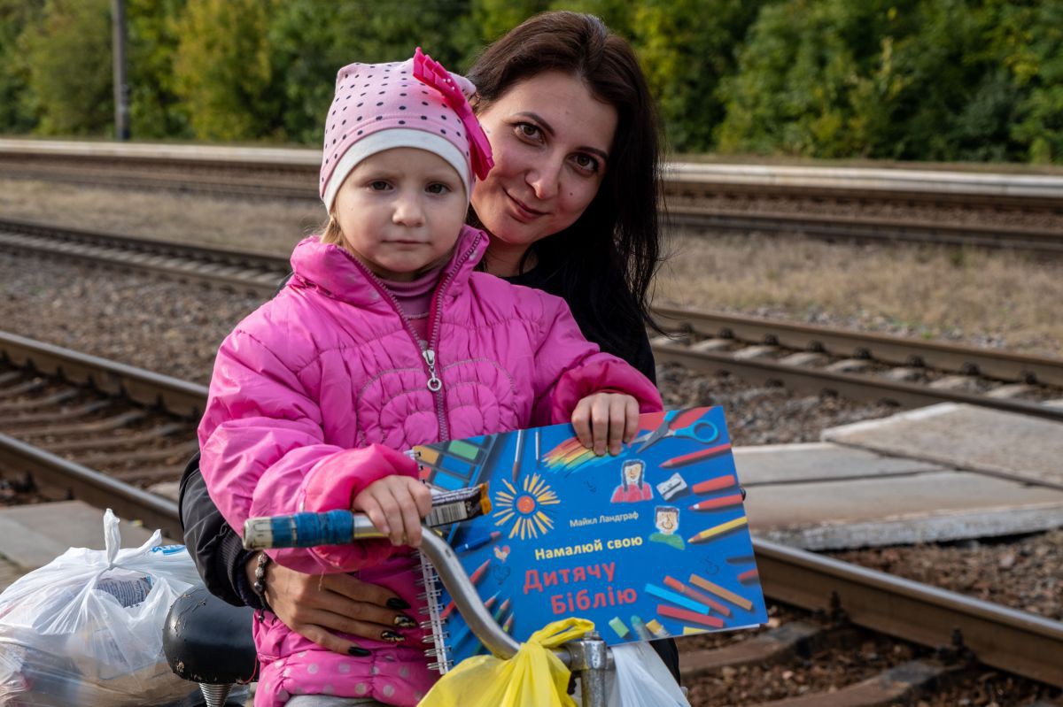 Eine junge Grau mit einem Kind in Daunenjacke auf dem Schoß. Das Kind hält eine Kinderbibel zum Selbstgestalten in der Hand.
