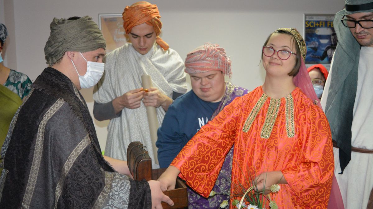 Alle erleben mit, wie Zachäus Jesus begegnet. Menschen verkleidet als biblische Figuren. Ein Mädchen in einem orangenem Kostüm greift in eine geöffnete Schatzkiste, die eine andere Person in Händen hält.