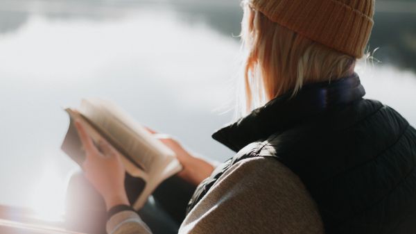 Eine Frau mit Mütze liest am Ufer eines Sees die Bibel.